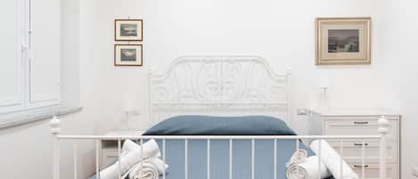 A neatly made bed with white linens, a blue mattress cover, and rolled towels on the side. There are two framed pictures on the wall, a white bedside table with a lamp, and a white nightstand with a drawer.