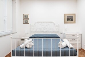 Ein ordentlich bezogenes Bett mit weißen Laken, einer blauen Matratzenauflage und aufgerollten Handtüchern. An der Wand hängen zwei gerahmte Bilder, ein weißer Nachttisch mit Lampe und ein weißer Beistelltisch mit Schublade.