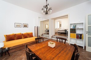 A modern living room with a wooden dining table, a sofa with orange cushions, a chandelier, and a bookshelf. - Maison Nunzia (Naples)