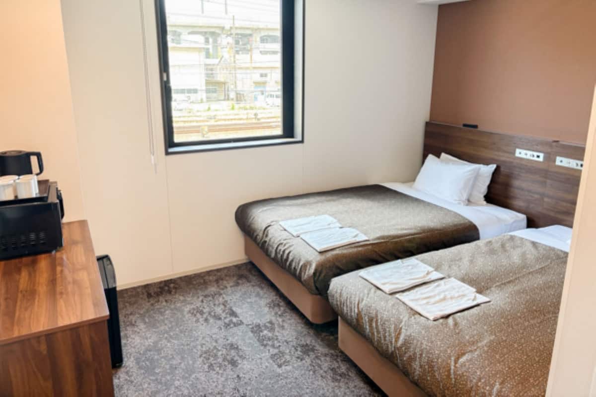 A hotel room with two beds, a desk, and a window with a view of buildings.