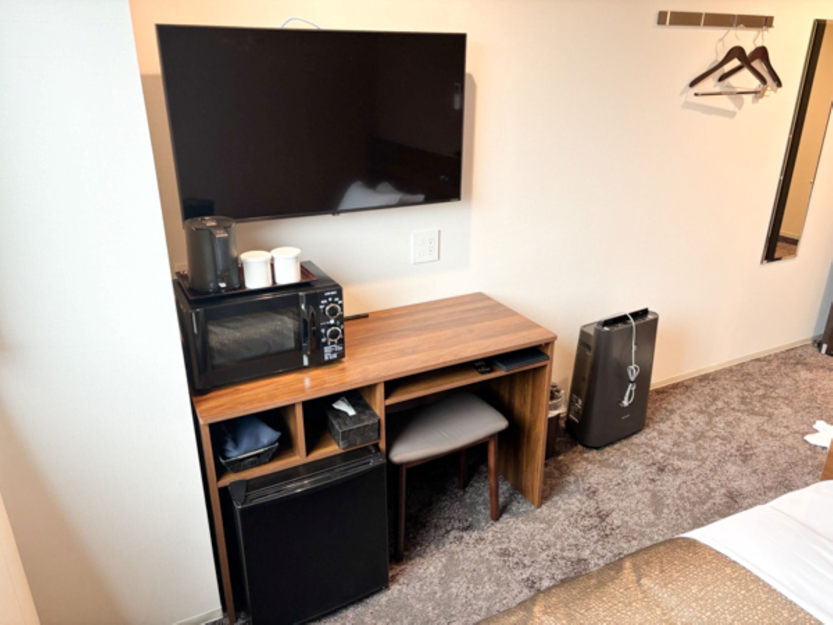 A compact hotel room with a wooden desk, a small refrigerator, a microwave, a TV mounted on the wall, and a suitcase.