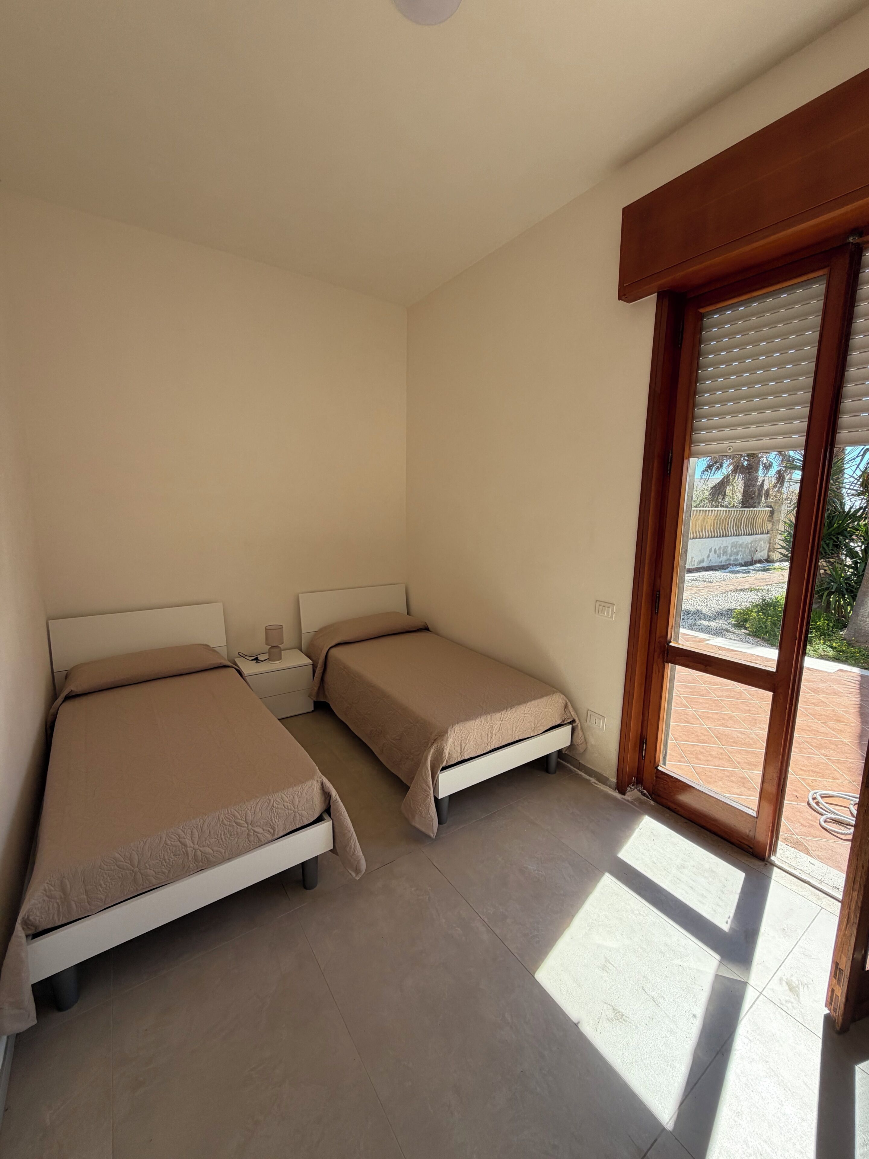 A room with two beds, a nightstand, a window with blinds, and a view of the outside.
