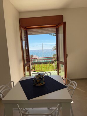 Quarto com mesa e cadeiras, vista para o mar através de uma porta aberta, e varanda com grade.