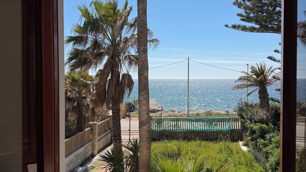 Una vista attraverso una finestra con cornice in legno, che dà su un balcone con ringhiera, una zona verde protetta da una rete e un cielo azzurro limpido.