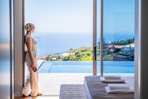 Eine Frau steht an einem großen Fenster mit Blick auf einen Pool und den Ozean.