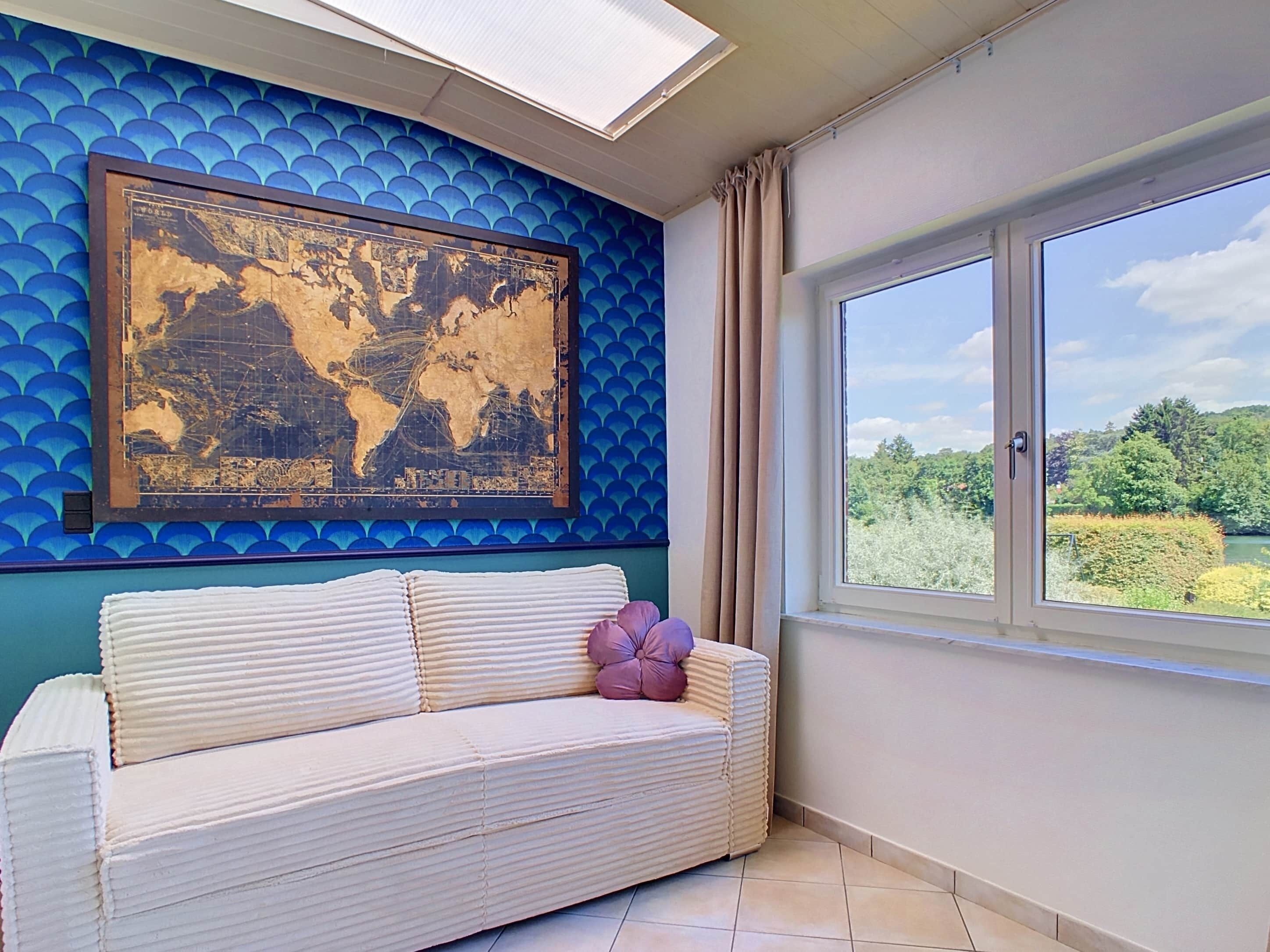 A room with a world map artwork, a sofa with cushions, a window with a view of greenery, and a skylight.