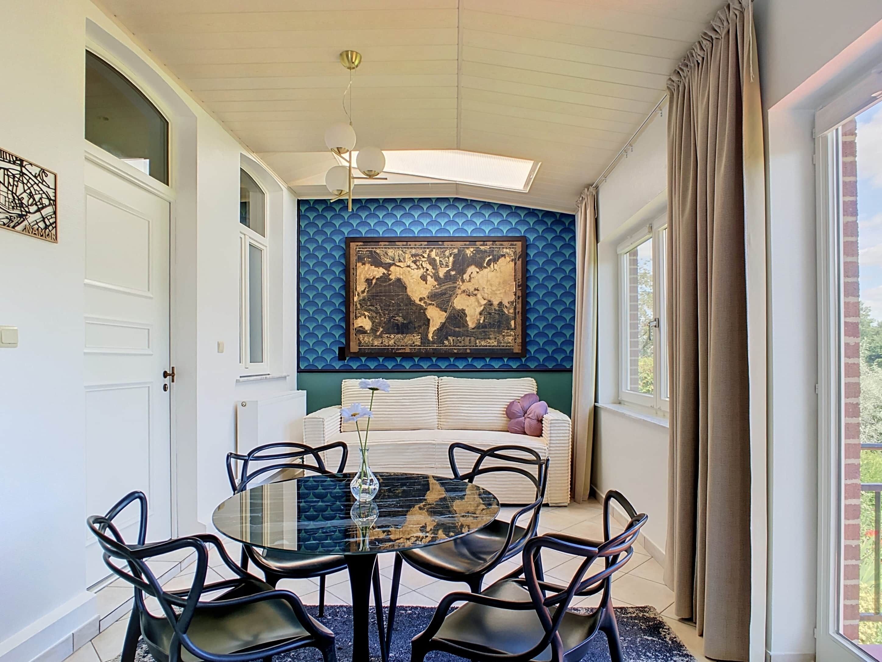 A dining area with a round glass table, black chairs, a world map on the wall, and a window with curtains.