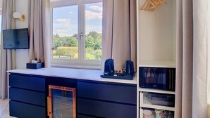 Een moderne kamer met een groot raam, een flatscreen televisie, een bureau met lades, een koffiezetapparaat en een kledingkast met houten hangers.