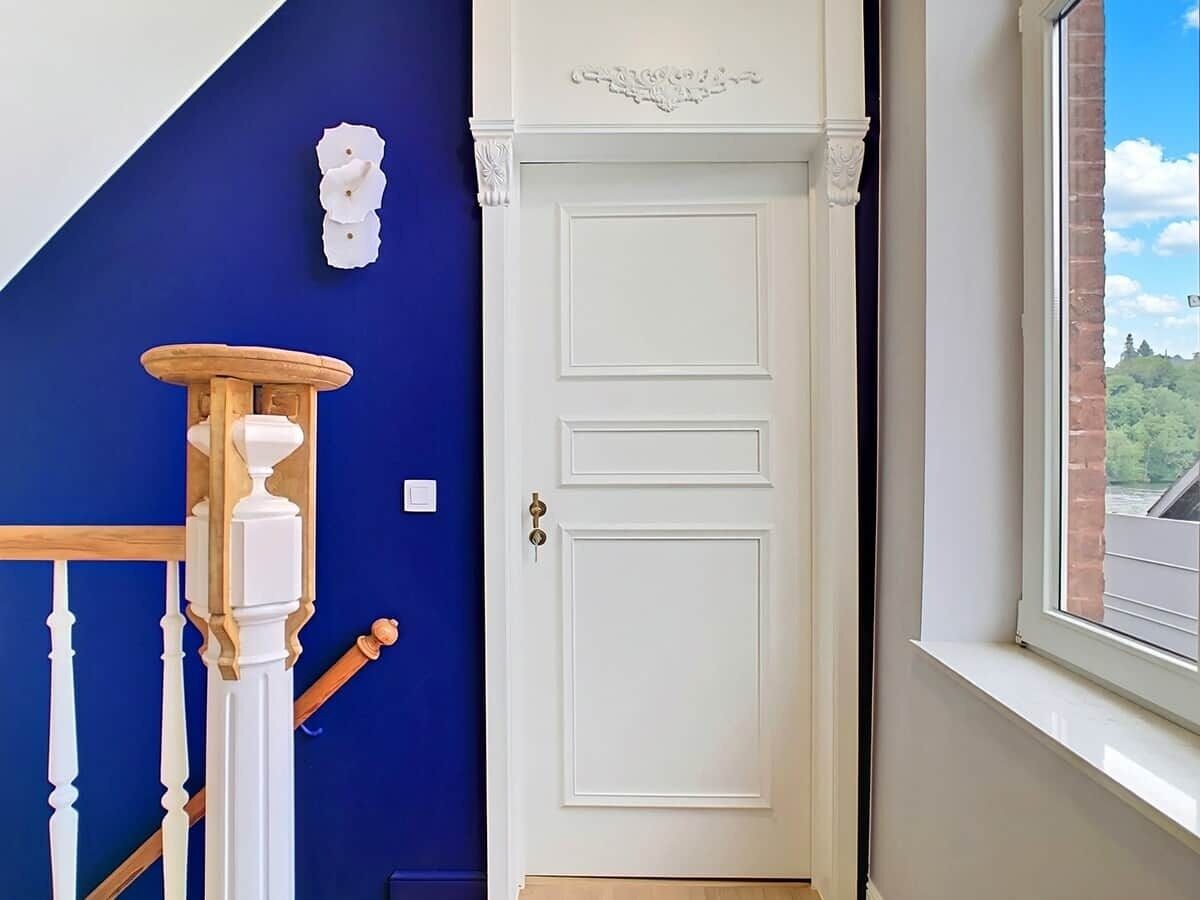 Une porte blanche avec une poignée en laiton, un mur bleu et une fenêtre donnant sur un paysage verdoyant.
