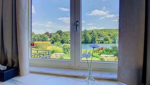 Een kamer met een groot raam, met uitzicht op een meer, begroeiing en een huis in de verte.