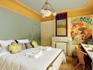 A hotel room with a bed, a desk, a chair, a radiator, and a wall with a mural.