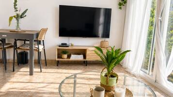 A living room with a flat-screen TV, a glass coffee table, a dining area with chairs, and a large window with curtains.