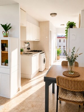 A modern kitchen with white cabinetry, a washing machine, and a dining area with wicker chairs and a table. - Cala Pupi Terrace, Quiet, Cozy and Bright Accommodation (Calafell)
