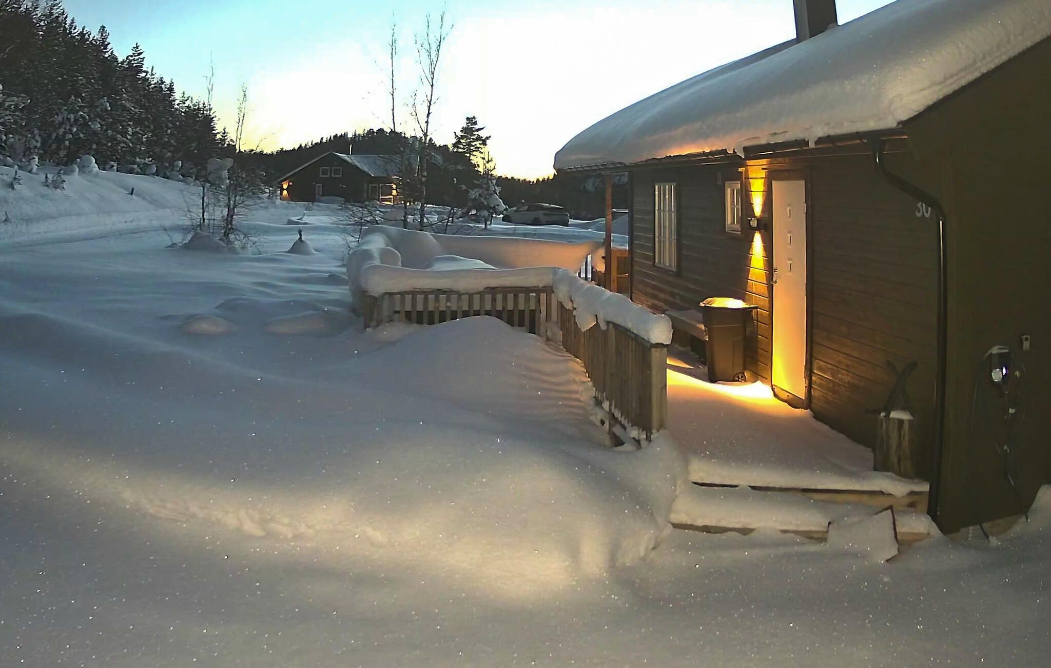 Une cabane en bois avec un porche enneigé et un intérieur éclairé, entourée d’un paysage hivernal.