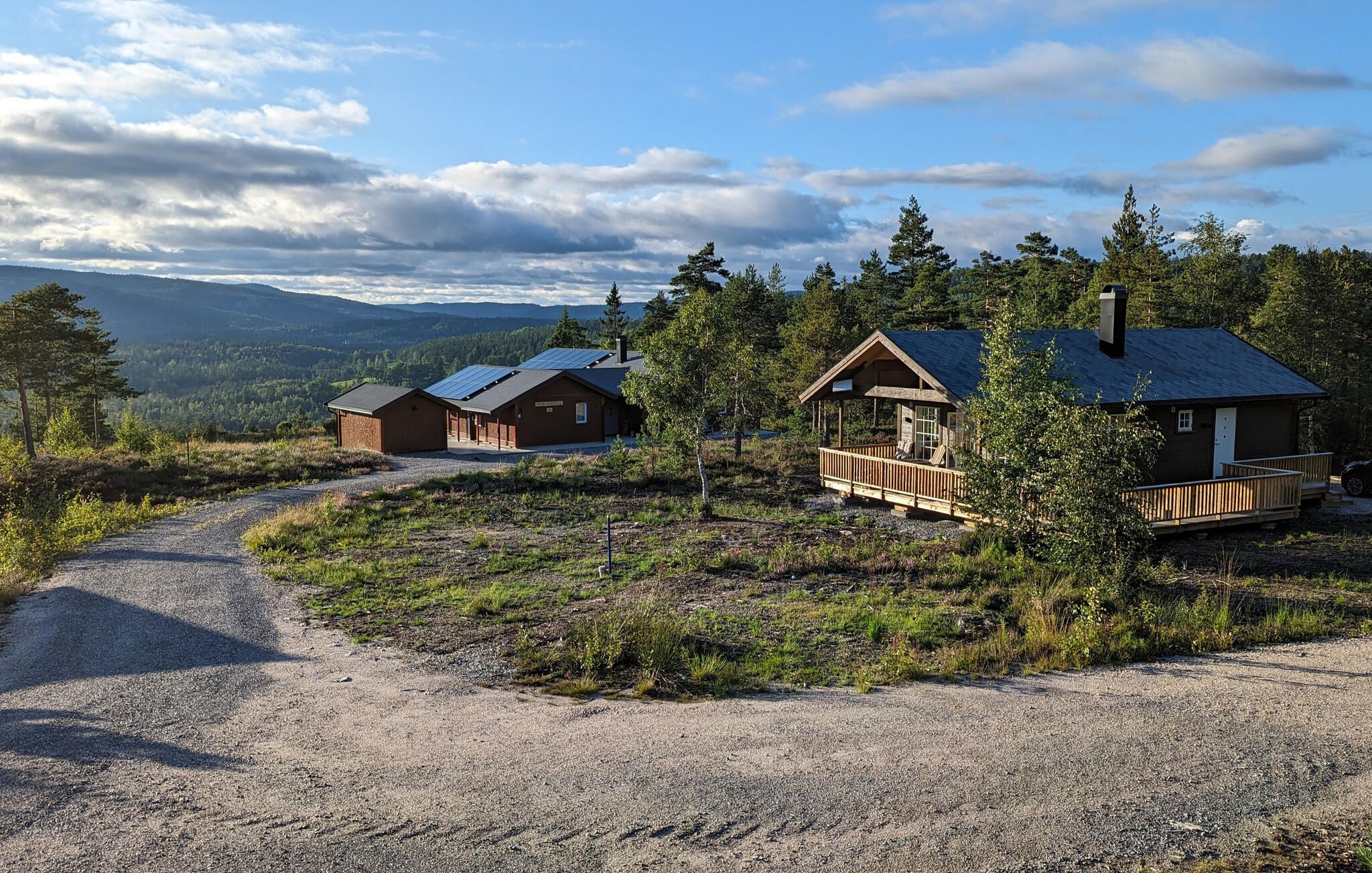 Uma cabana com um caminho de cascalho que a leva, rodeada por árvores e um cenário montanhoso.