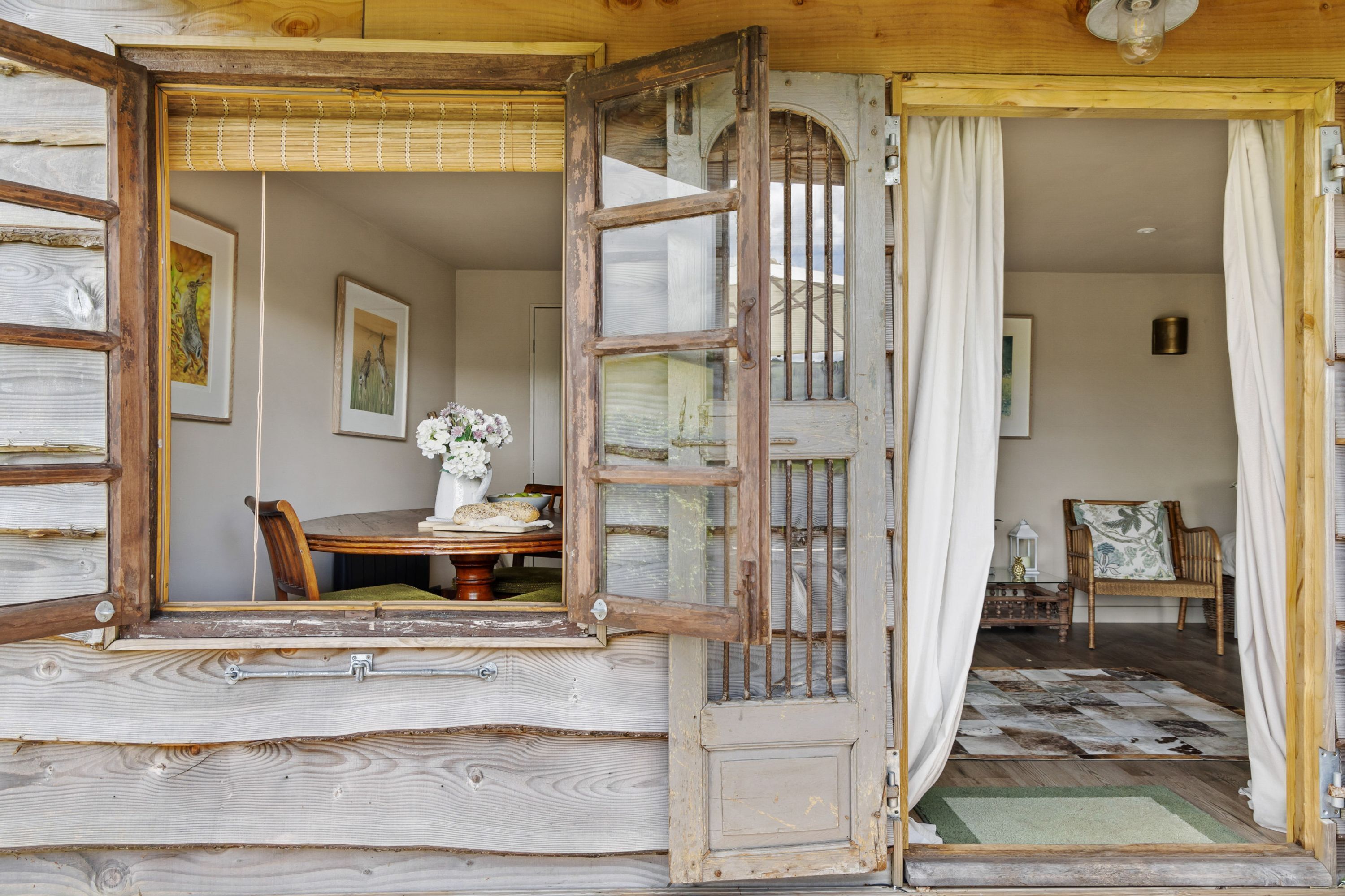 Une porte en bois ouverte donne sur une pièce où se trouvent une table à manger, des chaises et un vase rempli de fleurs.