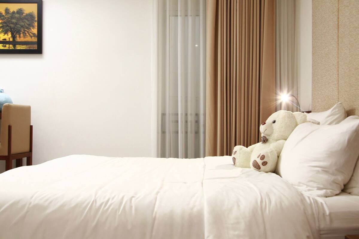 A hotel room with a bed, a chair, a window with curtains, and a teddy bear on the bed.
