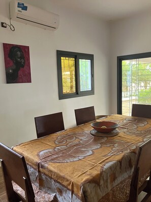 A dining area with a table covered in a patterned tablecloth, six chairs, a wall-mounted air conditioner, a window with a metal grill, and a framed artwork on the wall.
