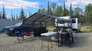 A camping setup with a covered table, chairs, a grill, and a trailer. - Kenai River RV at Fish Camp + Processing (Soldotna)