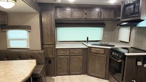 A compact RV kitchen with a stove, sink, and microwave, featuring wooden cabinets and a window with blinds. - Kenai River RV at Fish Camp + Processing (Soldotna)