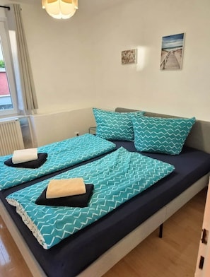 Ein Bett mit türkisfarben gemusterten Bettbezügen und Kissen, ein Fenster mit Vorhängen und ein Wandbild im Strandthema.