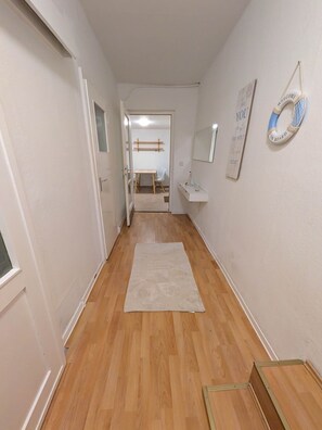 A hallway with a wooden floor, a white rug, a door leading to a room, a small table with a chair, a wall-mounted mirror, and a welcome sign. - Cozy "Koje" Vacation Rental with a Private Garden (Krummin)