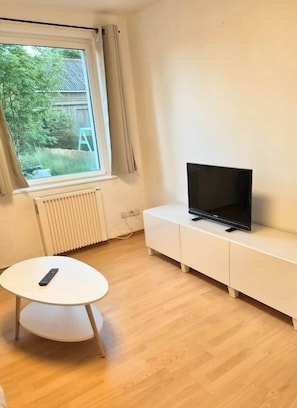 Ein Zimmer mit einem weißen Schrank, einem Fernseher darauf, einem kleinen runden Tisch mit Fernbedienung, einem Fenster mit Vorhängen und einem Heizkörper.