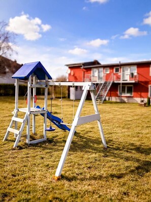 A playground with a swing set and slide, a blue-roofed structure, and a red building in the background.