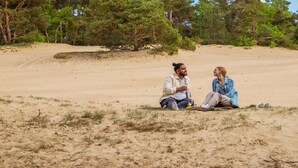 Twee personen zitten op het strand, met bomen op de achtergrond.
