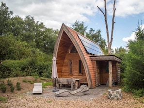 Eine Holzhütte mit geneigtem Dach, einer kleinen Veranda, einer Feuerstelle und einer Bank.