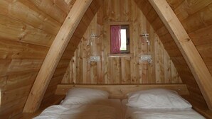 Een houten zolderkamer met een schuine plafond, een klein raam met een rood geruit gordijn, en een bed met witte beddengoed.