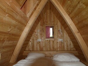 Ein Schlafzimmer im Dachgeschoss mit Holzbalken, einer kleinen, durch einen roten Karottenvorhang verhangenen Fensters und einem Bett mit weißer Bettwäsche.