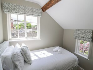 Un dormitorio con cama, dos ventanas adornadas con cortinas estampadas, vista a zonas verdes y techo inclinado.