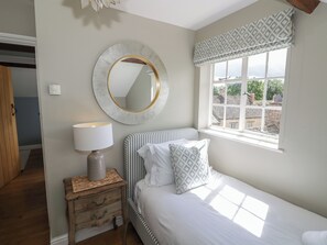 A bedroom with a bed, a bedside lamp, a round mirror, a window with patterned valance, and a view of buildings outside. - Tea Tree Cottage (Chipping Campden)