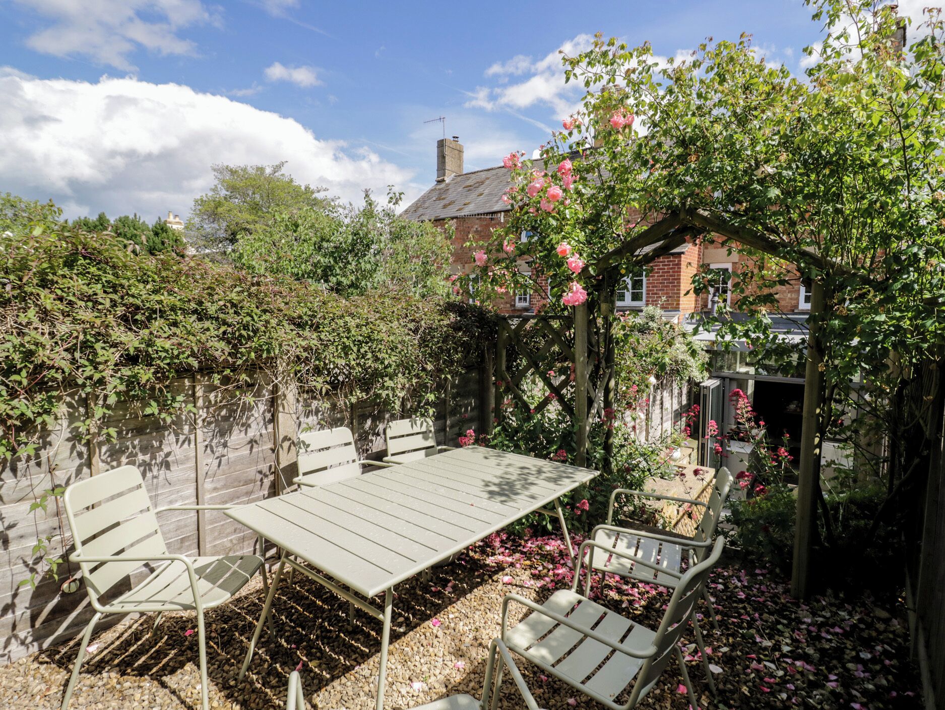 A garden patio with a table and chairs, surrounded by greenery and a brick building.