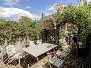 Une terrasse de jardin avec une table et des chaises, entourée de verdure et bordée d’un bâtiment en briques.