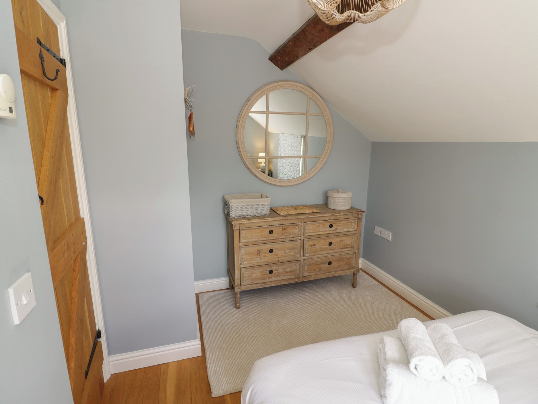 Ein Schlafzimmer mit einem Holzkoffer, einem runden Spiegel, einem Bett mit weißer Bettwäsche und einem Holzbalken an der Decke.