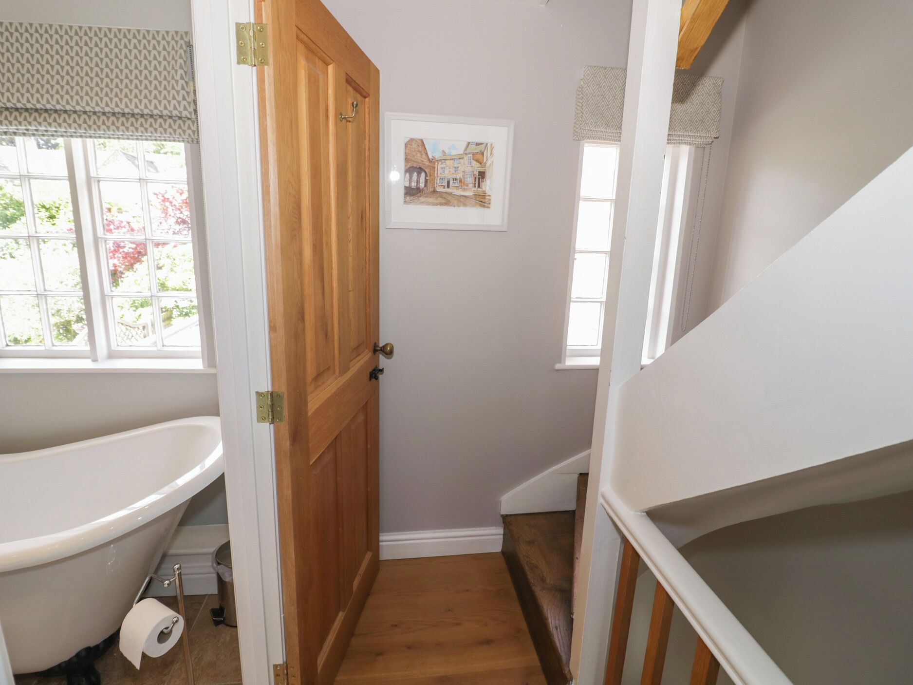 A narrow bathroom with a wooden door, a bathtub, and a window with blinds.