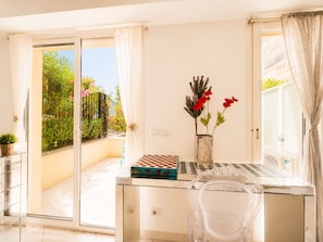 Ein Zimmer mit Glastisch, Schachbrett, einer Vase mit roten Blumen und Blick auf einen Garten durch eine Schiebetür.