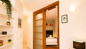 A hotel room with a sliding door, a bed, a small table with a lamp, and shelves with plants and a framed picture. - Beachfront Apartment in Menton (Menton)