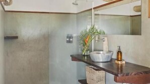 A bathroom with a concrete shower, wooden shelves with toiletries, a mirror, and a small plant. - Pousada Pais Tropical (Amontada)