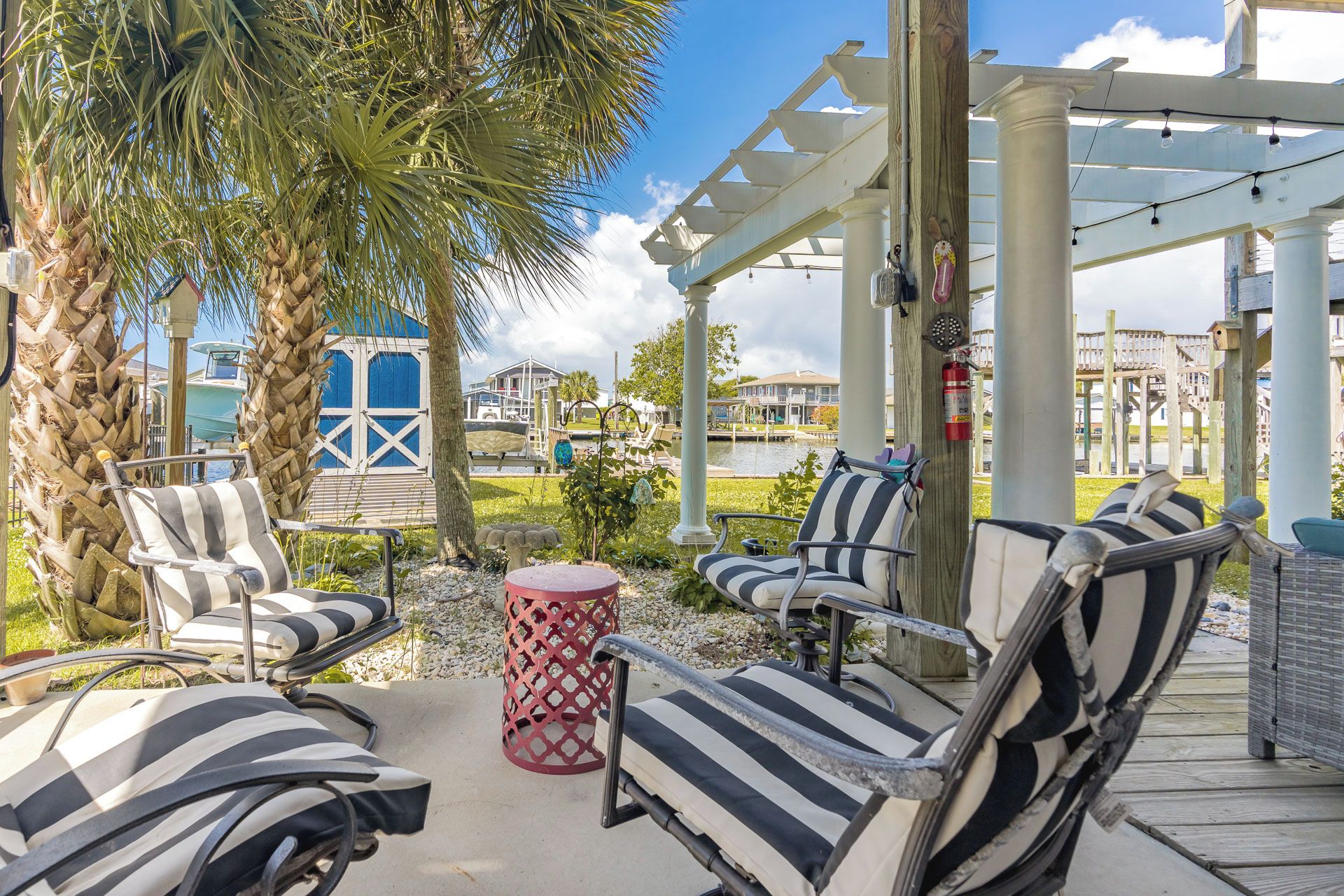 An outdoor seating area with striped chairs and a red table, overlooking a marina with boats and houses.