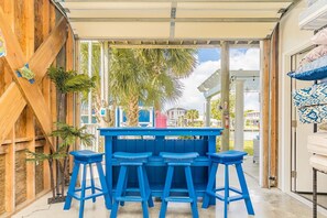 A covered outdoor area with a blue bar and stools, palm trees, and a view of a residential area. - Sunset Landing - 4 BR home on Canal - covered porch, private dock, pet friendly! (Surf City)