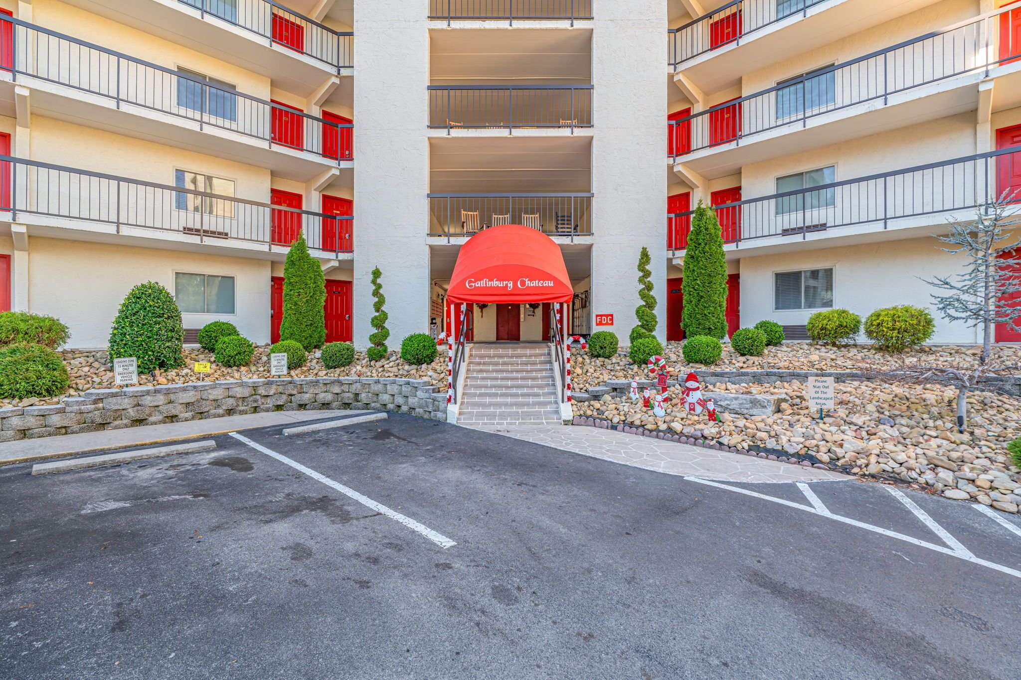 A multi-story hotel with red balconies, a central entrance with a red awning, and landscaped gardens with neatly trimmed bushes and a stone pathway.