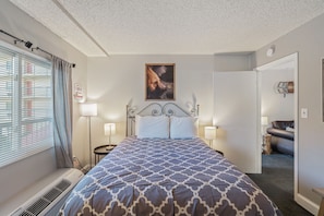 Ein Hotelzimmer mit einem großen Bett, einem Kopfteil mit einem gerahmten Bild, Nachttischlampen, einem Fenster mit Jalousien und einer Tür zu einem anderen Raum.