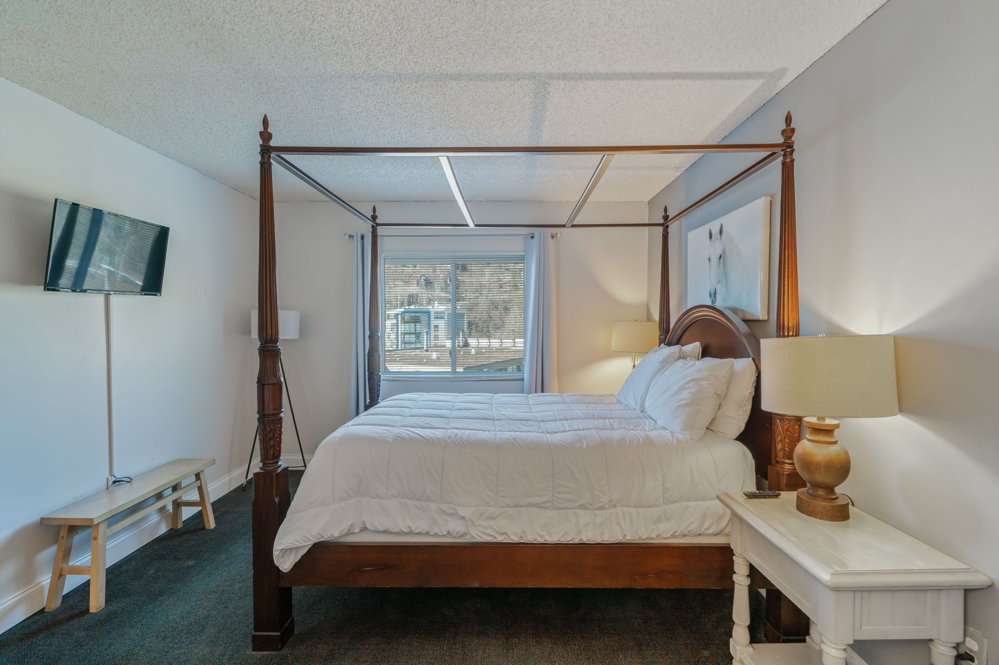 A four-poster bed with white bedding, a bedside table with a lamp, a TV mounted on the wall, and a window with blinds.