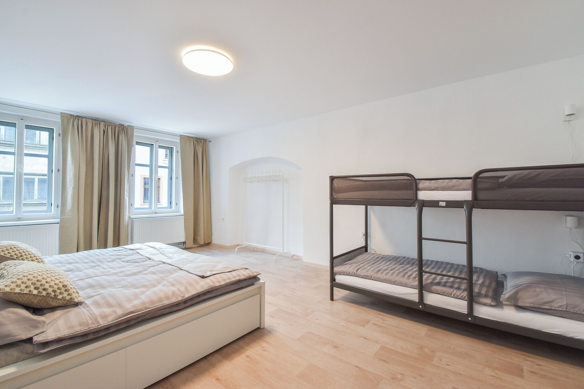 A bedroom with a bunk bed, a single bed, a window with curtains, and a radiator.