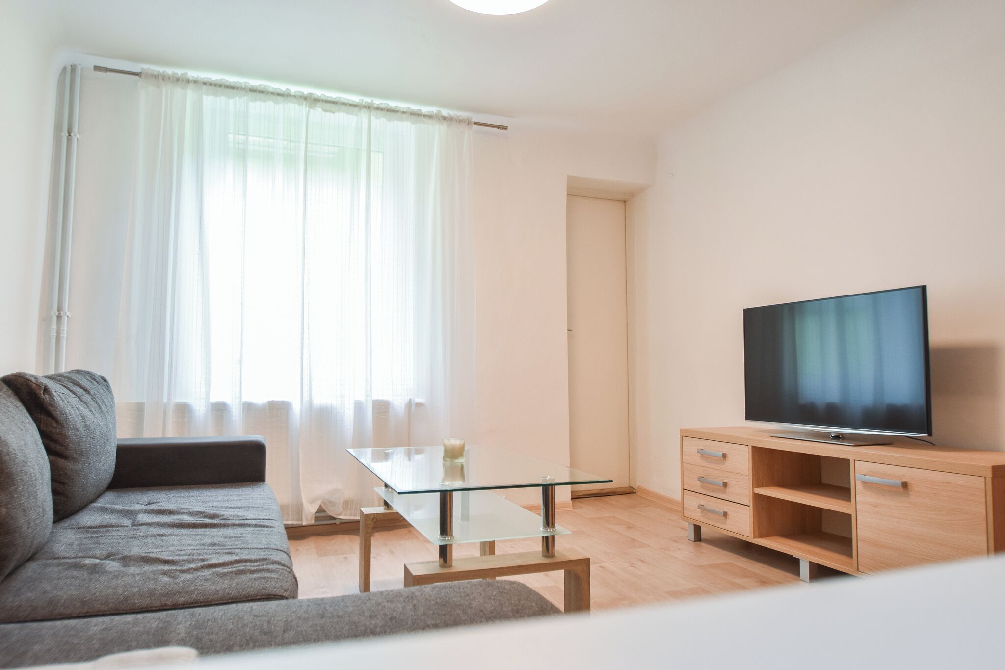 Una sala de estar moderna con un sofá, una mesa de centro de vidrio, un televisor sobre un mueble de madera y una ventana con cortinas translúcidas.