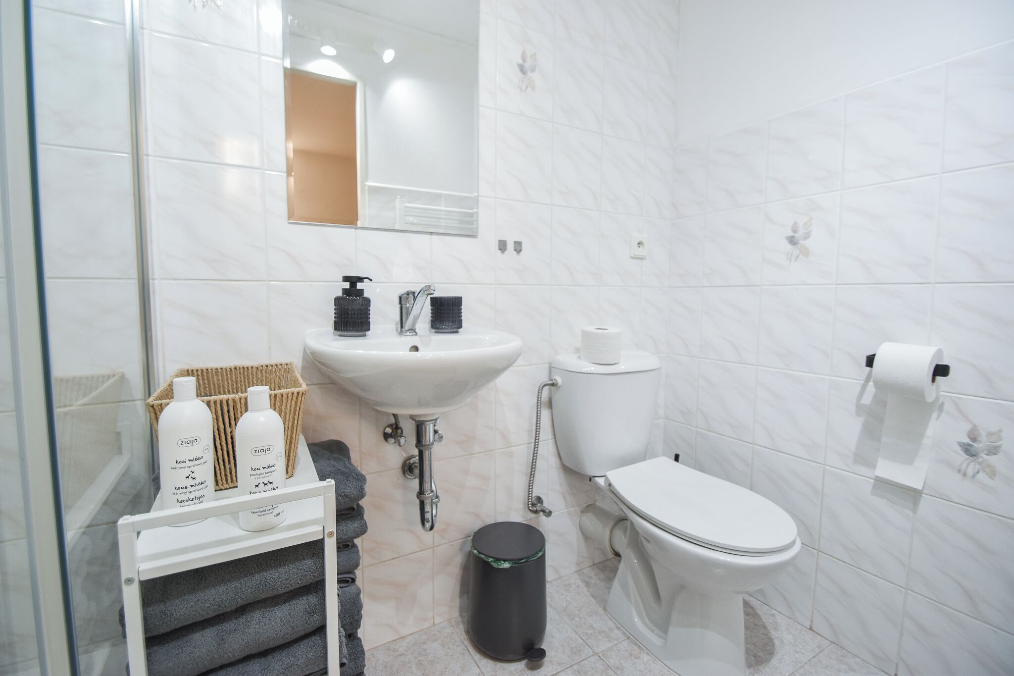 Une salle de bain moderne avec un lavabo, des toilettes et un miroir, tous de couleur blanche. On y trouve des articles de toilette sur une étagère et une poubelle.