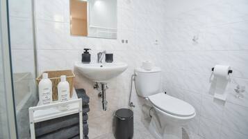 Une salle de bain moderne avec un lavabo, des toilettes et un miroir, tous de couleur blanche. On y trouve des articles de toilette sur une étagère et une poubelle.